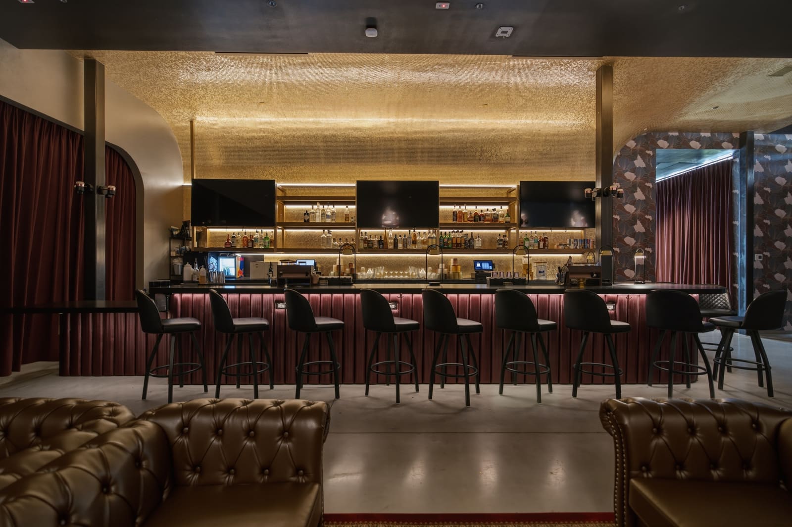 The lounge bar straight on: a row of stools along the red bar front, bottles lit on the back bar, screens above