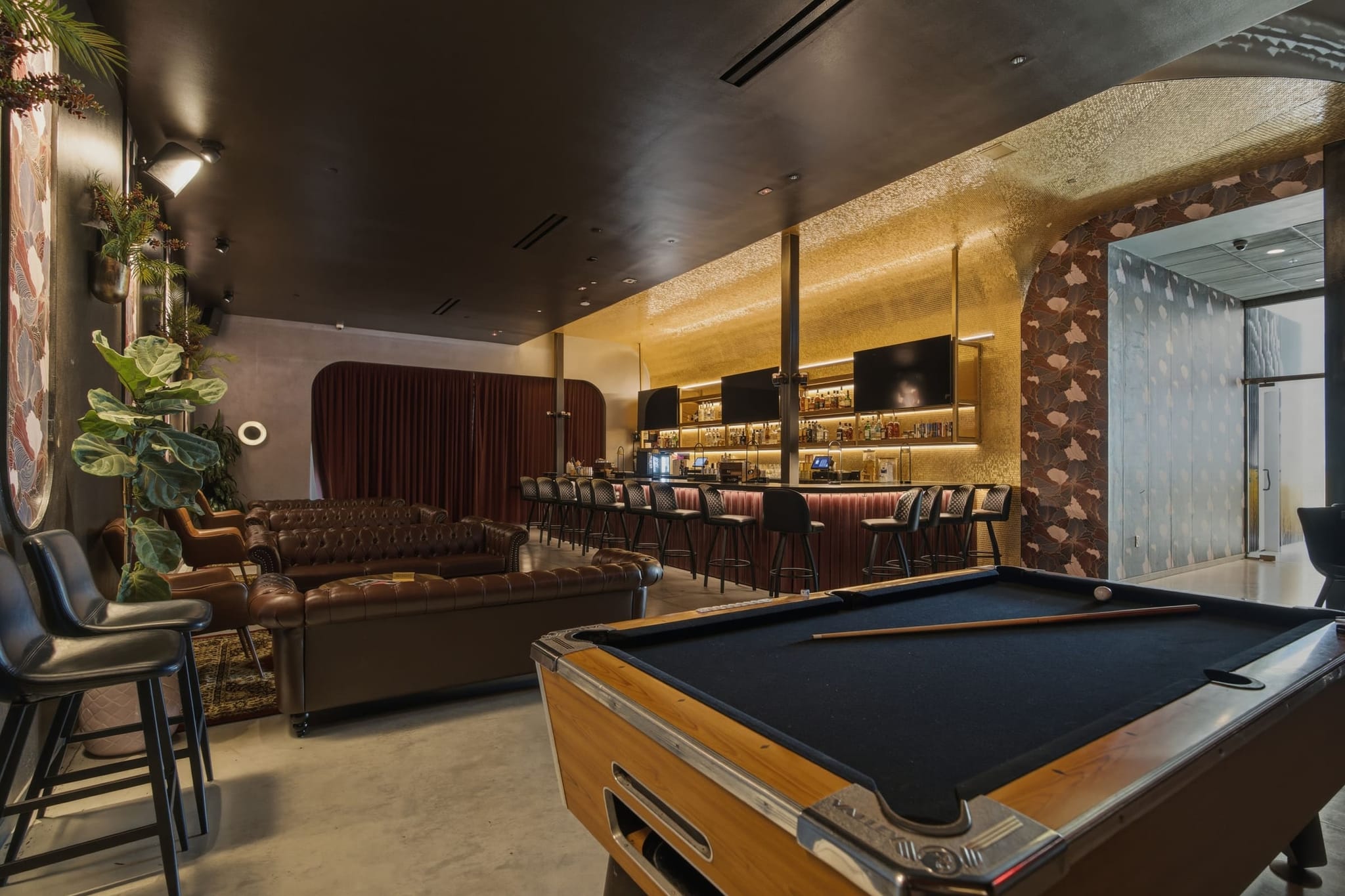 The Assembly Icehouse lounge, empty: the pool table in the foreground, leather couches on rugs, and the lit bar with screens along the back wall